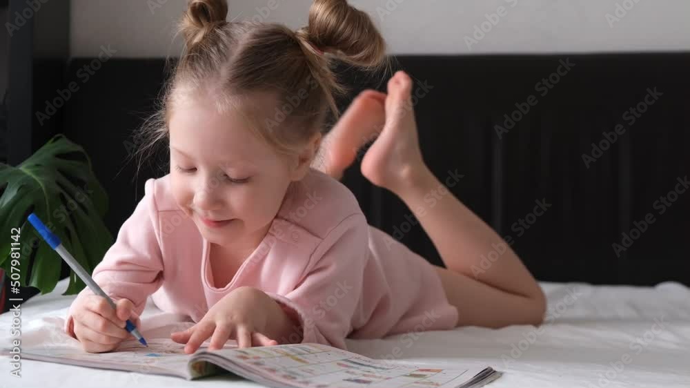 Portrait of a schoolgirl solves children crosswords on the bed. Logic games at home. Children solves crossword
