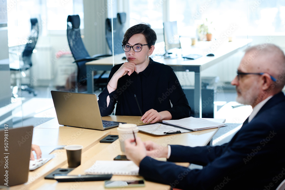 Serious young businesswoman sitting at table with laptop with pensive expression during meeting with her colleagues at office Serious young businesswoman sitting at table with laptop with pensive expression during meeting with her colleagues at office