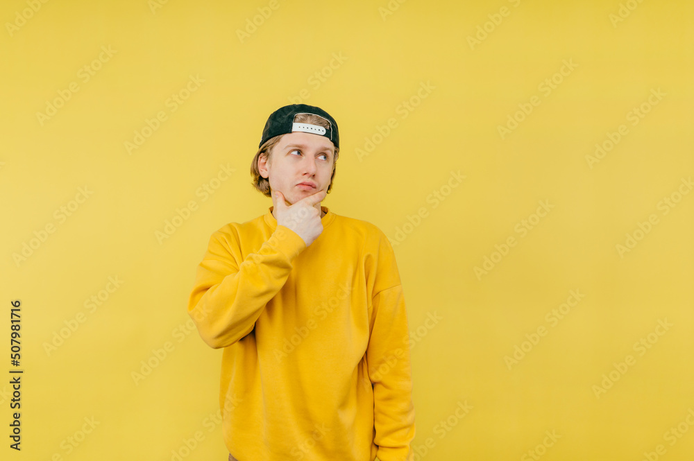 Portrait of a pensive guy in a sweatshirt and cap stands on a yellow background and looks away and thinks. Portrait of a pensive guy in a sweatshirt and cap stands on a yellow background and looks away and thinks.