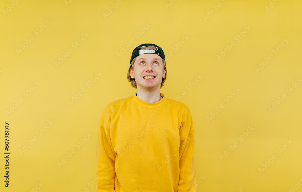 Positive young man in a cap and sweatshirt with a radiant face looking up on a yellow background Positive young man in a cap and sweatshirt with a radiant face looking up on a yellow background