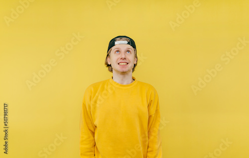 Positive young man in a cap and sweatshirt with a radiant face looking up on a yellow background