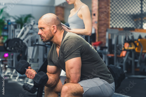 Profile indoor shot of a hairless bearded strong male bodybuilder staying focused during weightlifting, sitting on a bench in a nice well-equipped fitness space. High quality photo