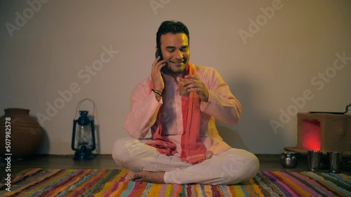 Medium shot of an attractive Indian villager talking on a phone while having tea. Village scene of a handsome middle-aged farmer busy over a phone call while drinking tea at home - lifestyle concept photo