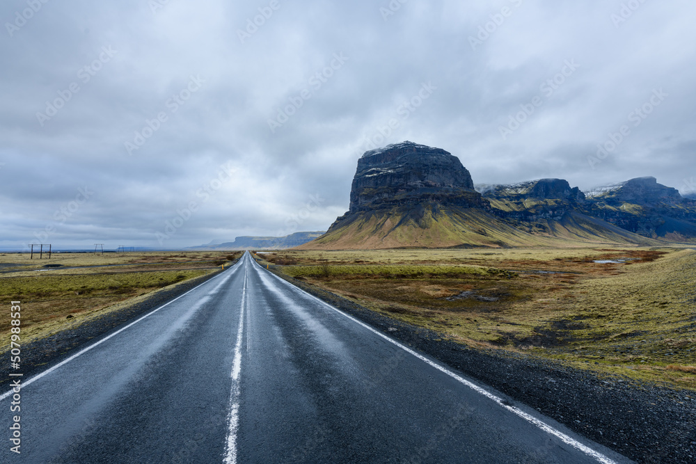 Straight road accross green barren scandinavian landscape with mountain ridge and peaks, cloudy sky