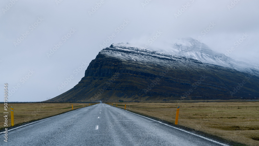 Straight road over autumn grass fields towards moody snow powdered scandinavian peak with sky grey and foggy