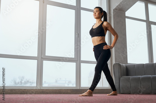 Side view of caucasian woman staying in position of yoga, looking straight while training at home. Slim female yogi with ponytail practicing yoga exercise in spacious studio. Concept of yoga practice.