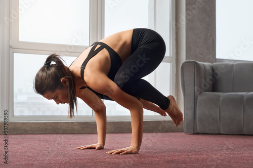 Side view of flexible woman twisting body in crow pose during yoga training at home. Slim female with ponytail standing in kakasana while practicing yoga in empty, light room. Concept of yoga. photo