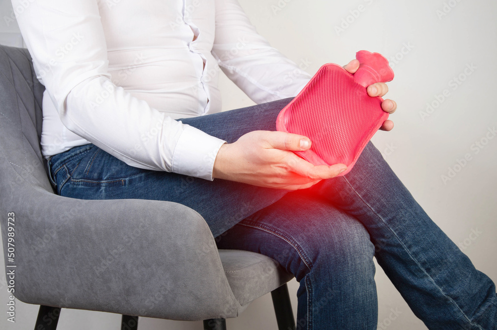 A male office worker holds a heating pad with hot water near the knee ...