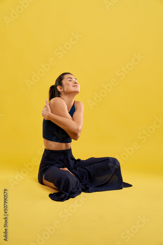 Fototapeta Naklejka Na Ścianę i Meble -  Young woman in spacious wear sitting in self hug pose, enjoying exercise in studio. Side view of happy girl wrapping arms around body in hug, isolated on orange background. Concept of relaxation.