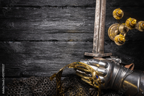 Photo Ancient golden crown, armor gloves and knight sword in the light of burning candle on the wooden table flat lay background with copy space