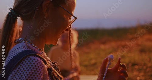 Back view of teenage girl with glasses eating roasted marshmellow, sitting over bonfire at summer campsite. Cheerful group of kids cooking meal on fire, resting in countryside. Concept of camping. 