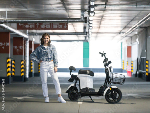 a girl posing with an electric scooter stands in a covered parking lot on a running board with an alarm
