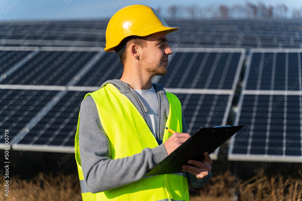 Successful worker man wearing protective clothes holding clip board taking notes solar panels source energy. Green energy concept Successful worker man wearing protective clothes holding clip board taking notes solar panels source energy. Green energy concept