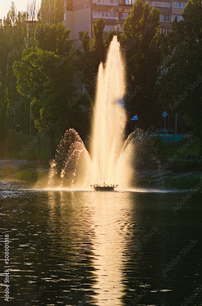 Picturesque landscape view of pond with fountain during sunset. Fountain in the city channel.  Buildings of famous Rusanivka neighborhood in the background. Kyiv, Ukraine