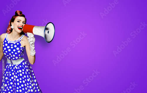 Portrait image of purple haired woman holding mega phone, shout advertising something. Girl in blue pin up style dress with mega phone loudspeaker. Isolated on violet background. Beauty model.