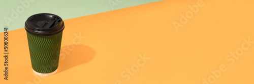 Banner with green paper coffee cup with black plastic lid over orange and green background with copy space. Sustainable food packaging concept