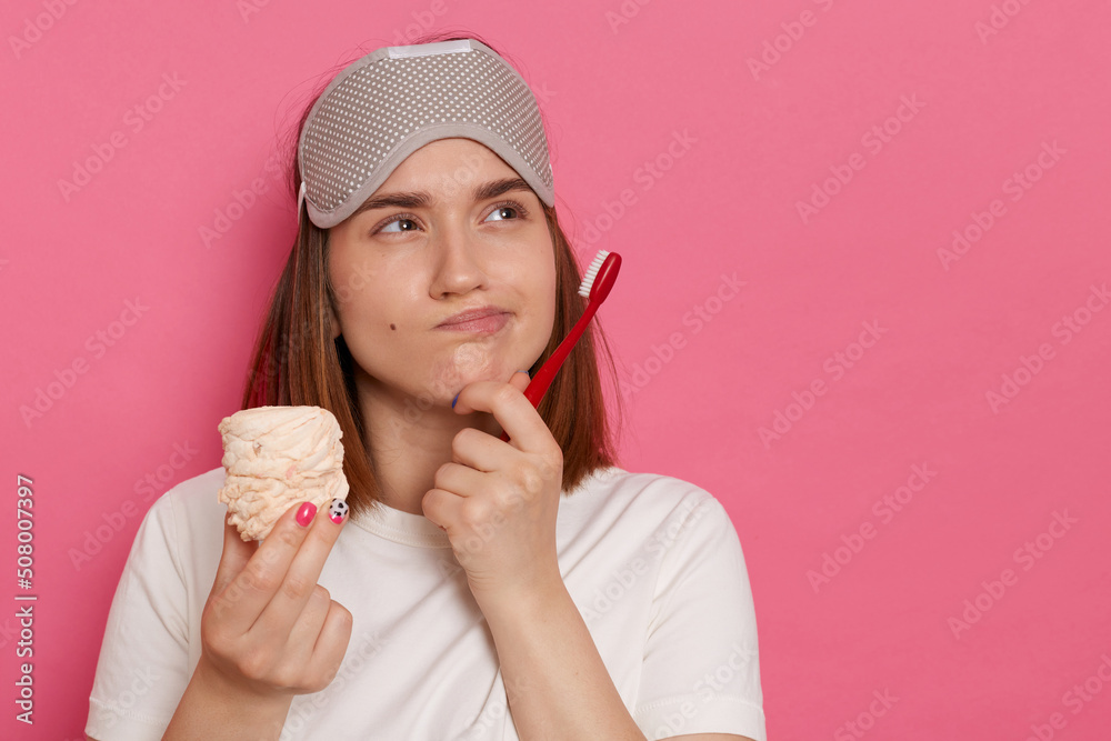 Indoor shot of sad pensive thoughtful woman with blindfold on her forehead, holding marshmallow and toothbrush, looking away, being deep in thoughts, wearing white casual style t shirt. Indoor shot of sad pensive thoughtful woman with blindfold on her forehead, holding marshmallow and toothbrush, looking away, being deep in thoughts, wearing white casual style t shirt.