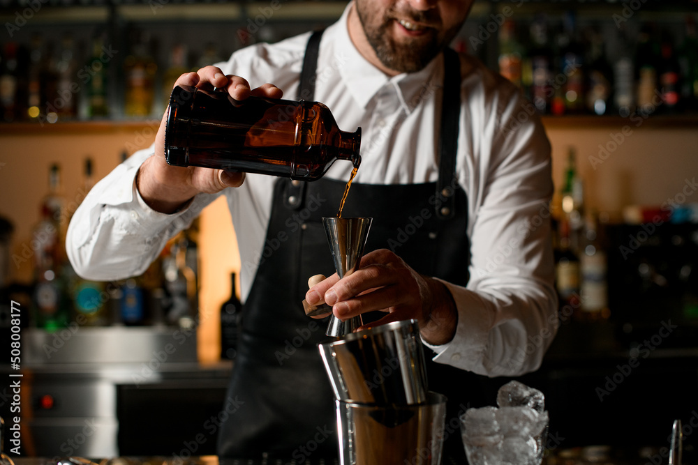 Close-up view of male bartender hands holding bottle and pours beverage into steel jigger Close-up view of male bartender hands holding bottle and pours beverage into steel jigger