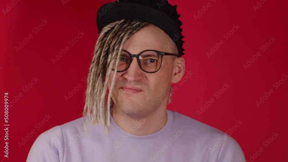 Young handsome man in black cap, glasses nodding head with disagreement. Stylish hipster with blonde dreadlocks showing gesture of protest with head on red background. Young handsome man in black cap, glasses nodding head with disagreement. Stylish hipster with blonde dreadlocks showing gesture of protest with head on red background.