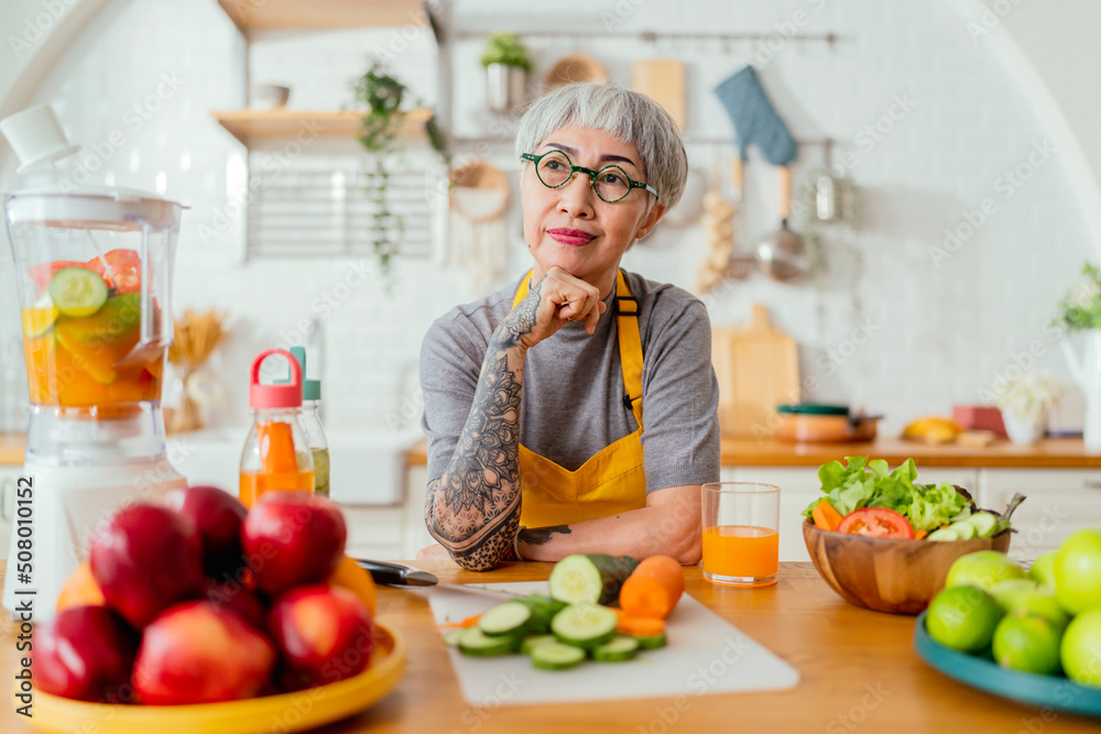 Mature smiling tattoo woman eating salad, fruits and vegetables. Attractive mature woman with fresh green fruit salad at home. Senior woman apron standing in the kitchen counter relaxing in house