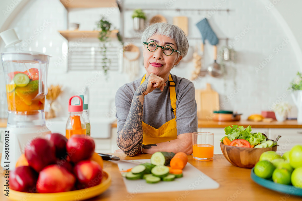 Mature smiling tattoo woman eating salad, fruits and vegetables. Attractive mature woman with fresh green fruit salad at home. Senior woman apron standing in the kitchen counter relaxing in house Mature smiling tattoo woman eating salad, fruits and vegetables. Attractive mature woman with fresh green fruit salad at home. Senior woman apron standing in the kitchen counter relaxing in house