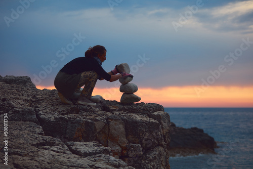 Canvas Print Silhouette of a woman balancing rocks and stones on the ocean sea coast at sunset sunrise time
