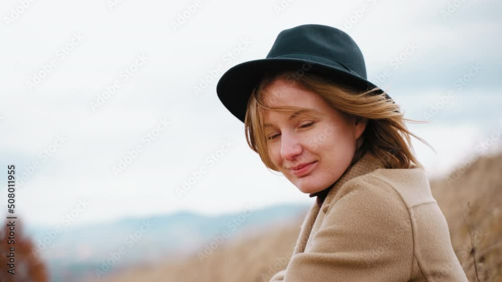 Portrait of beautiful smiling girl looking into camera blinks eyes. Woman in hat on head in open air in windy weather on background of mountains and sky. Lightness at soul, spiritual calm and freedom Portrait of beautiful smiling girl looking into camera blinks eyes. Woman in hat on head in open air in windy weather on background of mountains and sky. Lightness at soul, spiritual calm and freedom
