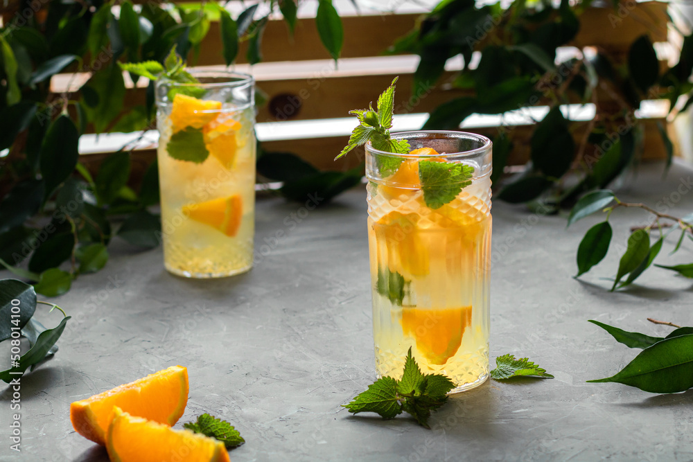 Two glasses with a refreshing orange drink. A type of lemonade. Mineral water with pieces of fresh orange and mint. Detox, diet. Behind the wooden lattice, entwined with greenery. Two glasses with a refreshing orange drink. A type of lemonade. Mineral water with pieces of fresh orange and mint. Detox, diet. Behind the wooden lattice, entwined with greenery.