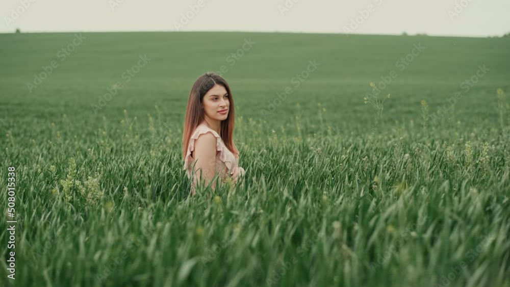 Young pregnant woman with flowing hair in a pink summer dress sitting among a green field with wheat. In the background a blue sky, a woman looking away Young pregnant woman with flowing hair in a pink summer dress sitting among a green field with wheat. In the background a blue sky, a woman looking away