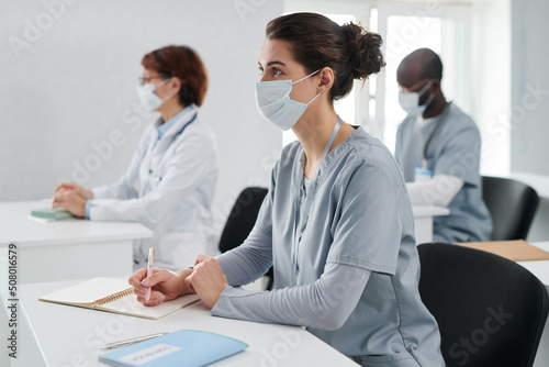 Young woman in mask sitting at desk with notebook and listening to speaker at medical training together with her colleague in background