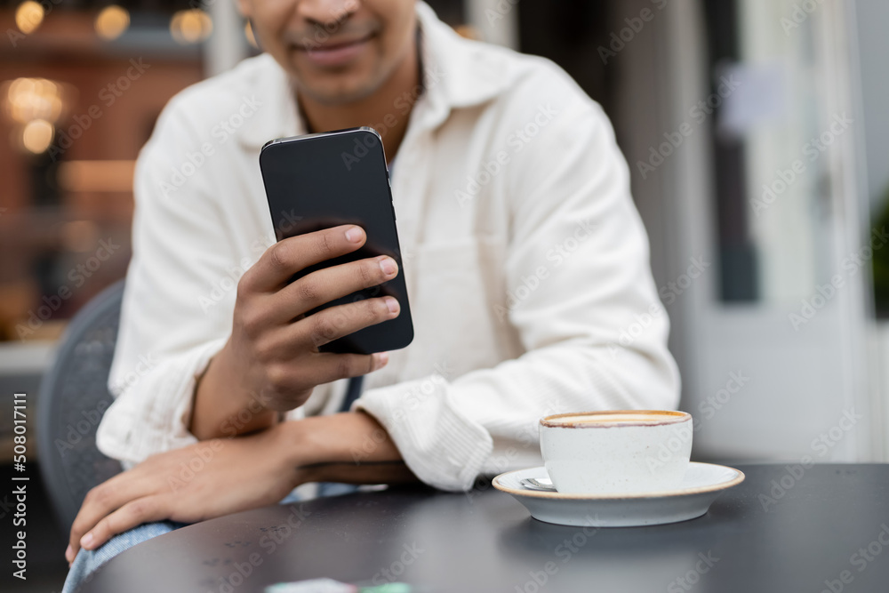 cropped view of blurred african american man texting on smartphone near cup of cappuccino on table on cafe terrace. cropped view of blurred african american man texting on smartphone near cup of cappuccino on table on cafe terrace.