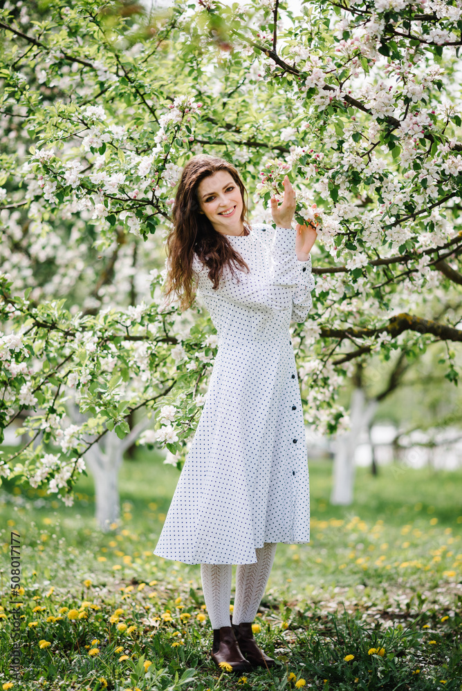 Portrait of adorable girl in beautiful dress walk on the green lawn in spring garden. Woman on the background of blooming apple and cherry trees.
