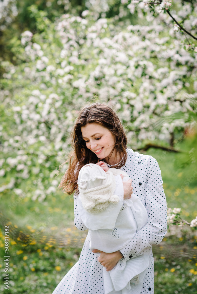 Mom holding baby girl, daughter in beautiful dresses walking on the green lawn in blooming apple blossom garden. Happy family concept. Mom holding baby girl, daughter in beautiful dresses walking on the green lawn in blooming apple blossom garden. Happy family concept.