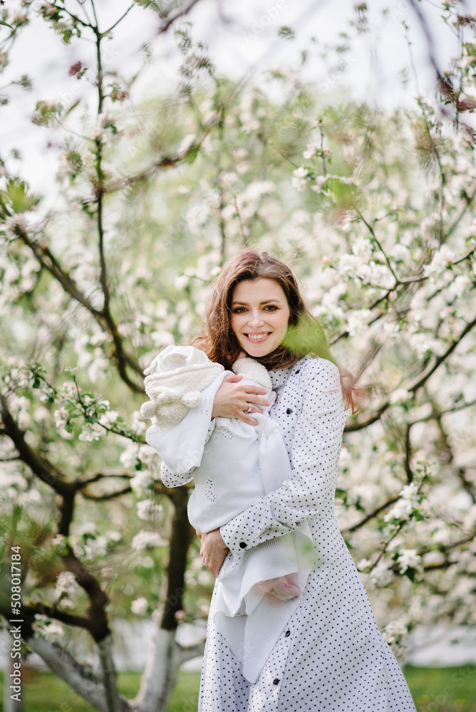 Mom holding baby girl, daughter in beautiful dresses walking on the green lawn in blooming apple blossom garden. Happy family concept. Mom holding baby girl, daughter in beautiful dresses walking on the green lawn in blooming apple blossom garden. Happy family concept.
