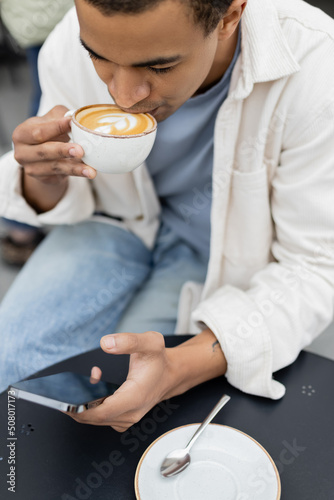 high angle view of african american man texting on smartphone and drinking cappuccino on cafe terrace.