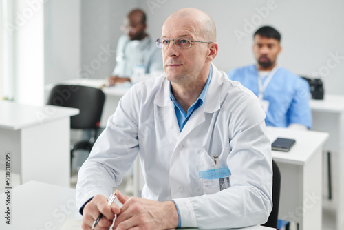 Mature male doctor in eyeglasses and white coat sitting at desk listening to lecture with serious expression