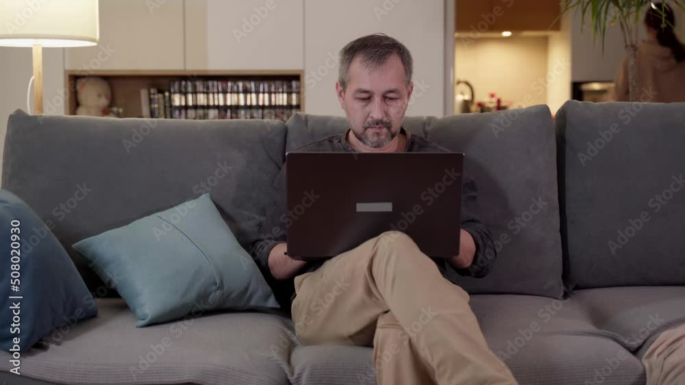 In the evening, a man with a laptop sits on the couch. Remote work at home. In the evening, a man with a laptop sits on the couch. Remote work at home.