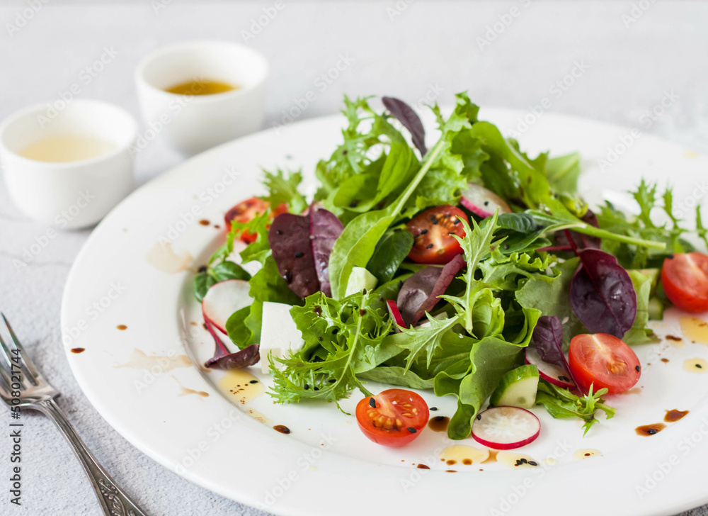 Green salad of fresh lettuce leaves, cherry tomatoes, feta cheese and assorted seeds with a sauce of honey, olive oil and balsamic vinegar on a flat plate on a stone table. Healthy vegan food.