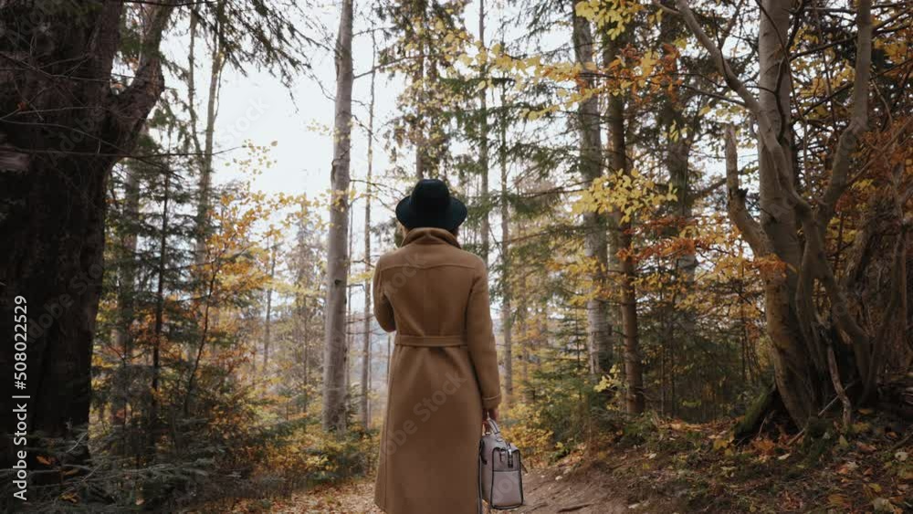 A lady in a coat and hat stands on a path in a fabulous picturesque forest and holds a briefcase backpack. Woman enjoys nature. Girl among autumn trees with bright leaves. Calm atmosphere A lady in a coat and hat stands on a path in a fabulous picturesque forest and holds a briefcase backpack. Woman enjoys nature. Girl among autumn trees with bright leaves. Calm atmosphere