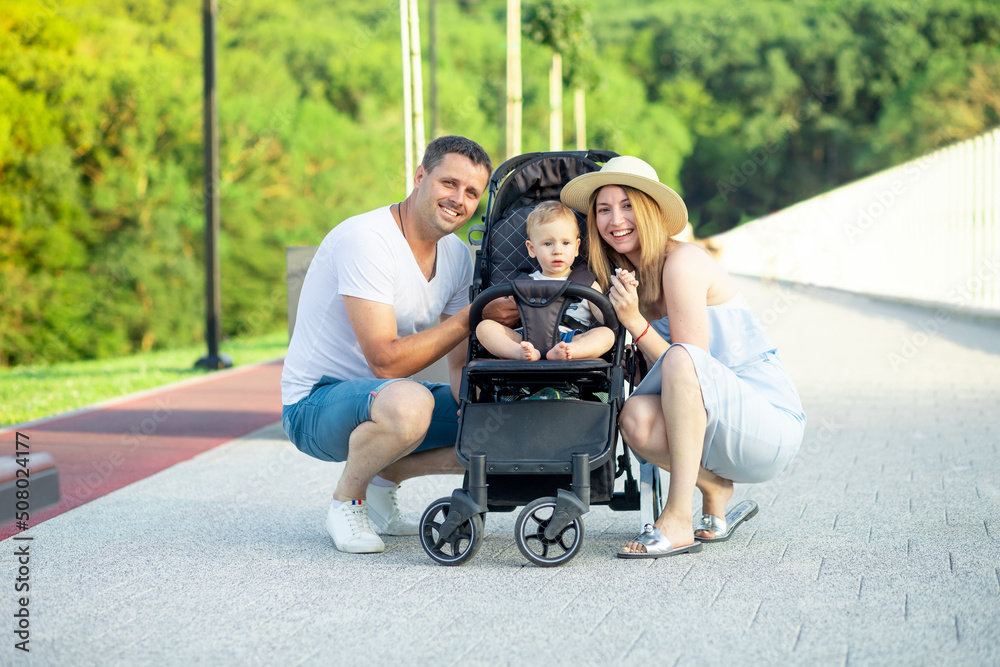 happy young parents mom and dad are walking with a baby in a stroller in the park in a hat in the summer in the setting sun and smiling having fun happy young parents mom and dad are walking with a baby in a stroller in the park in a hat in the summer in the setting sun and smiling having fun