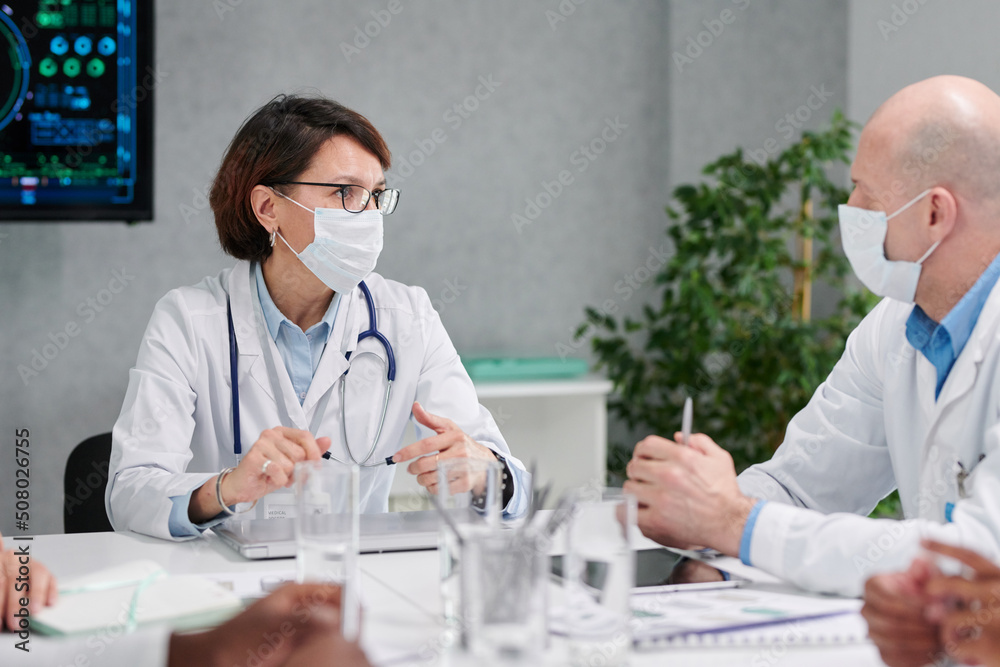 Two colleagues in masks and white coats discussing together at table during meeting at hospital Two colleagues in masks and white coats discussing together at table during meeting at hospital