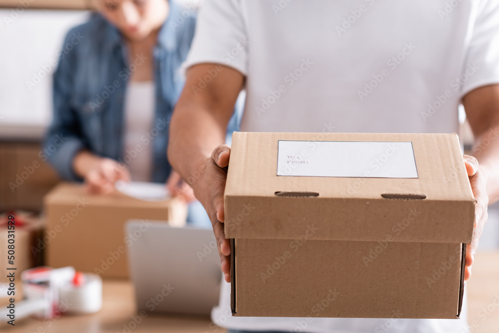 Cropped view of african american seller holding box with shipping label near blurred colleague in online web store. Cropped view of african american seller holding box with shipping label near blurred colleague in online web store.