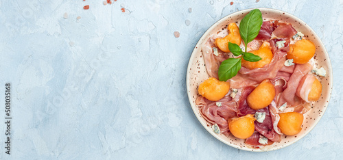 Fototapeta Naklejka Na Ścianę i Meble -  Summer appetizer with sweet melon and prosciutto and basil. Antipasti, Traditional Spanish and Italian appetizer. Top view on white stone table, Long banner format. top view