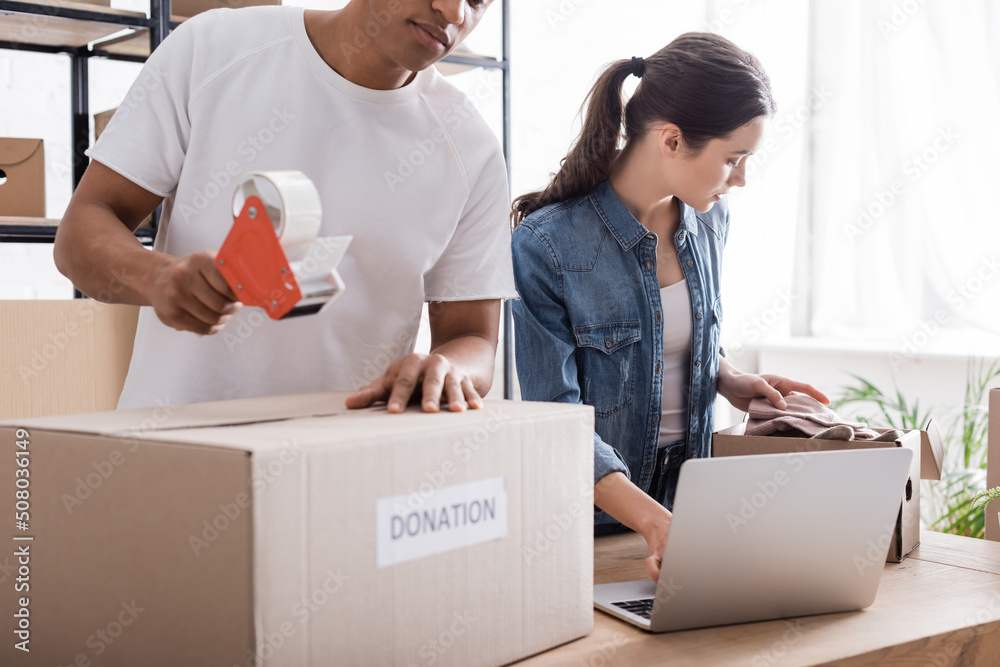 Multiethnic sellers packaging boxes with donation lettering near laptop in online web store. Multiethnic sellers packaging boxes with donation lettering near laptop in online web store.
