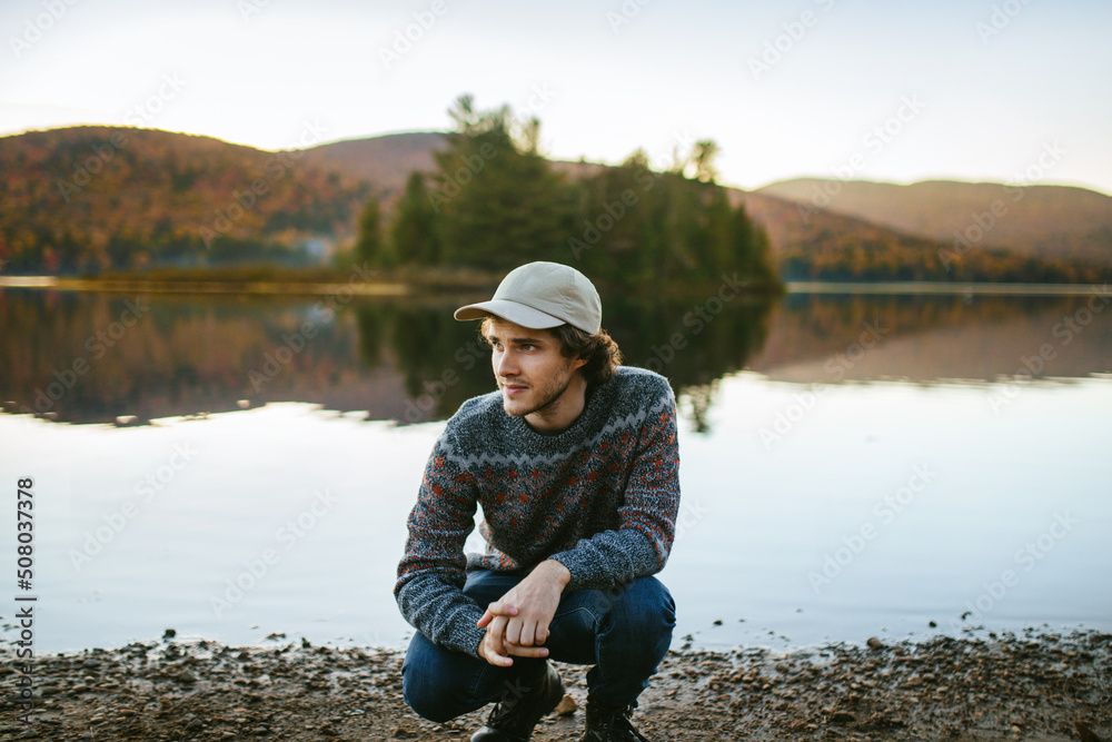 Stylish traveling guy sitting on haunches at lakeside on autumn day Stylish traveling guy sitting on haunches at lakeside on autumn day