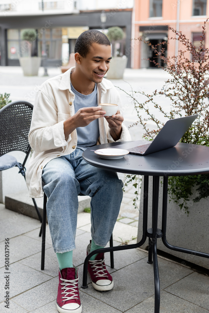 cheerful african american freelancer drinking coffee and using laptop on cafe terrace.