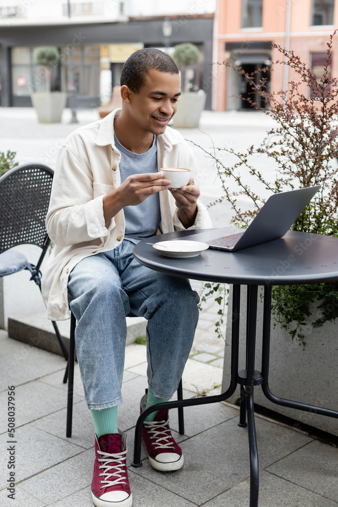 cheerful african american freelancer drinking coffee and using laptop on cafe terrace. cheerful african american freelancer drinking coffee and using laptop on cafe terrace.