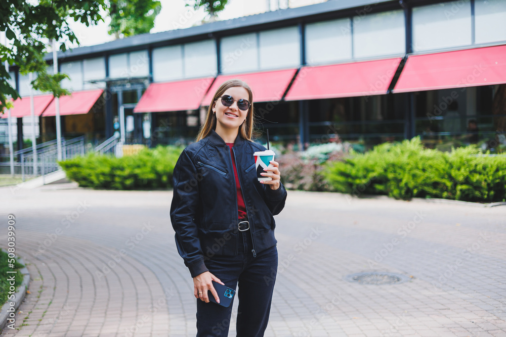 Beautiful and young girl in sunglasses writes a message on a cell phone and drinking coffee outdoors Beautiful and young girl in sunglasses writes a message on a cell phone and drinking coffee outdoors