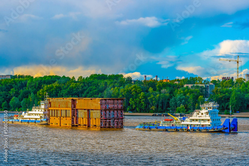 A tugboat pulling a heavy cargo barge with a container along the river carrying elements for the construction of the bridge and fuel.