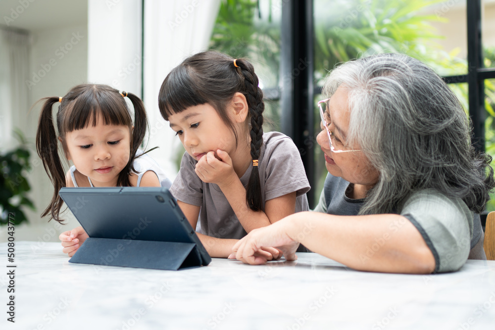 Asian Grandmother with her two grandchildren having fun and playing education games online with a digital tablet at home in the living room. Concept of online education and caring from parents.